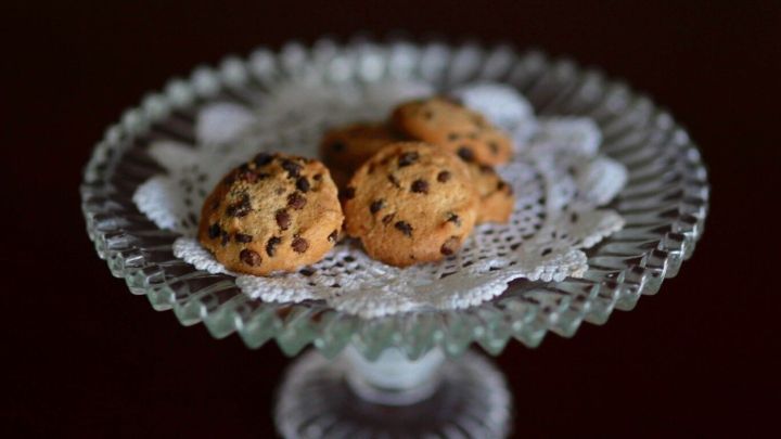 Auf einem eleganten Glasstil steht ein Teller mit knusprigen Mandel-Cookies, die mit Schokost&uuml;ckchen verziert sind.