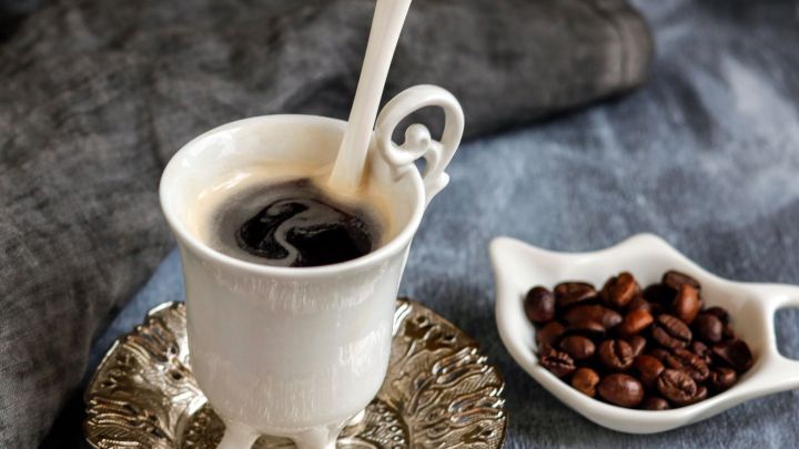 Eine elegante Tasse Kaffee steht auf einem filigranen Untersetzer, w&auml;hrend eine kleine Schale mit Kaffeebohnen daneben liegt.