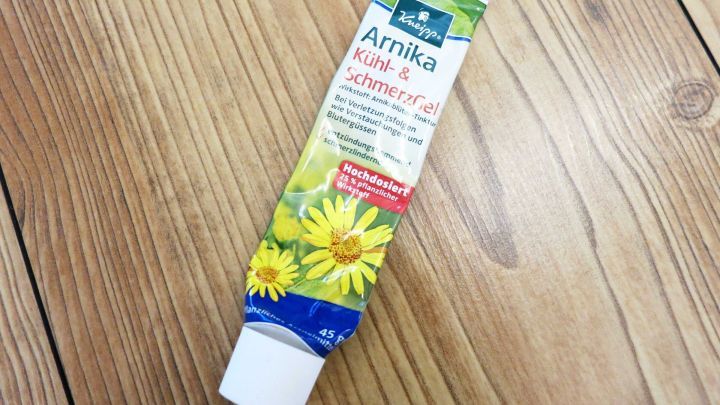 Eine Tube Arnikasalbe steht auf einem Holzfu&szlig;boden, bereit zur Anwendung gegen M&uuml;ckenstiche. Die Verpackung zeigt gelbe Blumen.