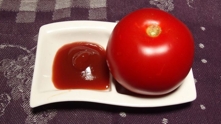 Eine rote Tomate liegt neben einer kleinen Schale mit Ketchup auf einem sch&ouml;nen Tisch mit Tuch.