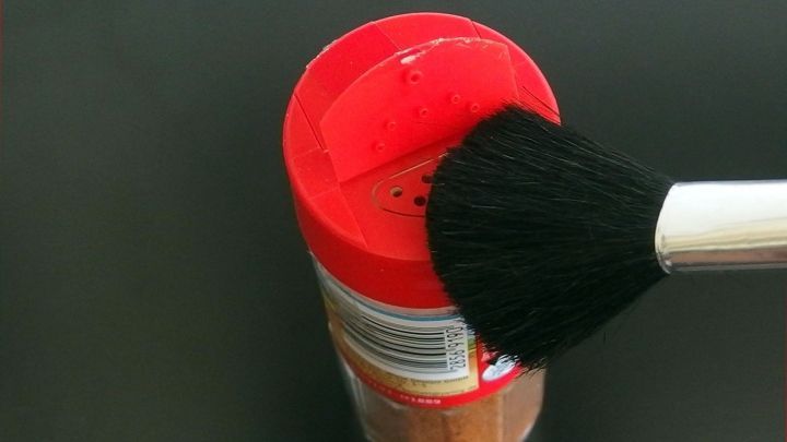 Ein roter Gew&uuml;rzstreuer wird mit einem Pinsel gereinigt, w&auml;hrend er auf einem schwarzen Tisch steht.