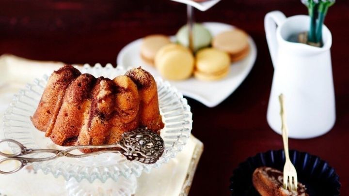 Ein Marmorkuchen steht auf einem eleganten Teller, w&auml;hrend er mit einer Gabel angeschnitten wird. Im Hintergrund befinden sich bunte Macarons und eine kleine Vase mit lila Blumen.