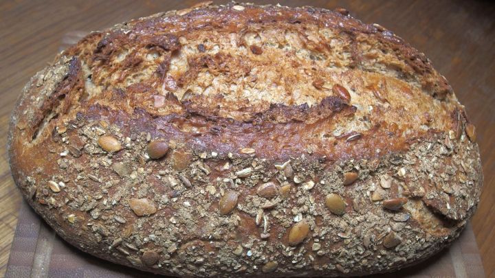 Ein frisches, krustiges Brot mit K&ouml;rnern liegt auf einem Holzbrett in einer gem&uuml;tlichen K&uuml;chenumgebung.
