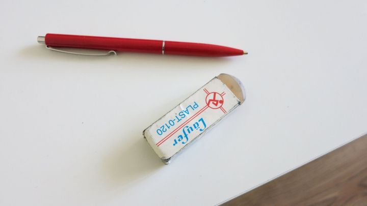Ein roter Kugelschreiber liegt neben einem Radiergummi auf einem wei&szlig;en Schreibtisch, bereit, Flecken zu entfernen.