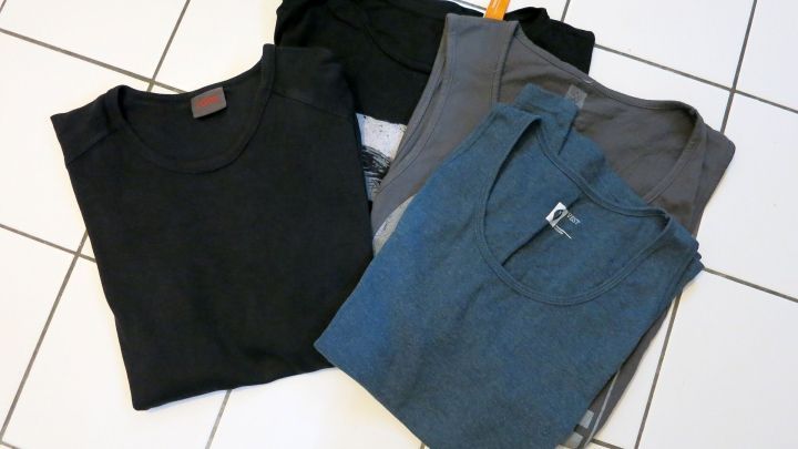 T-Shirts liegen gefaltet auf wei&szlig;en Fliesen, schwarz, grau, blau und mit Etiketten sichtbar.