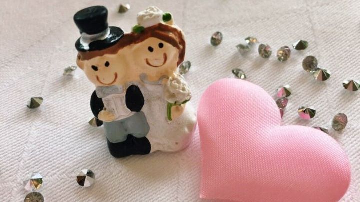Ein kleines Hochzeitspaar aus Keramik steht umgeben von glitzernden Streudekoren und einem rosa Herzen auf einem Tisch.