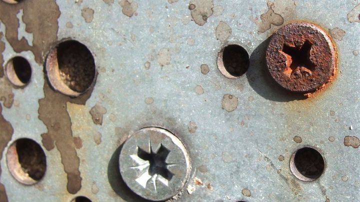 Eine rostige Schraube befindet sich neben einer sauberen, unversehrten Schraube auf einem metallenen Untergrund.