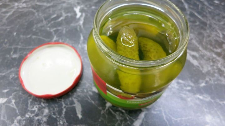 Ein Glas mit eingelegten Gurken steht offen auf einer dunklen, marmorierten Oberfl&auml;che, umgeben von einer entspannten K&uuml;chenatmosph&auml;re.