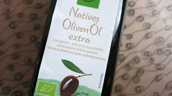 Eine Flasche mit nativem Oliven&ouml;l steht auf einem Tisch, umgeben von einem dezenten, gemusterten Hintergrund.