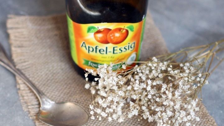 Eine Flasche Apfel-Essig steht auf einem rustikalen Untergrund, neben einer kleinen Schaufel und Trockenblumen.