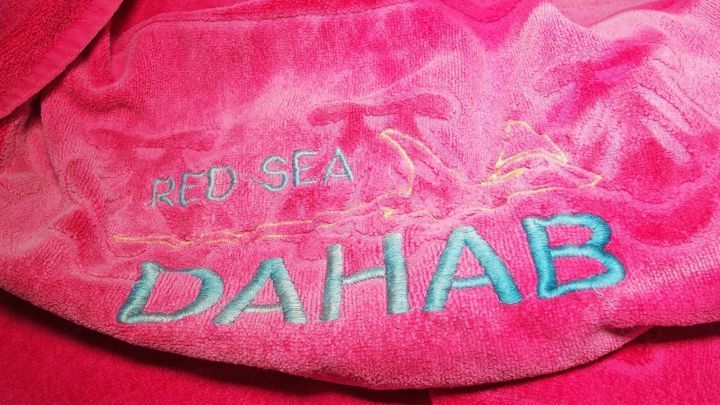 Ein pinkes Handtuch liegt auf einer Oberfl&auml;che. Darauf steht "RED SEA DAHAB" in blauen und wei&szlig;en Buchstaben, was auf einen Urlaubsort hinweist.