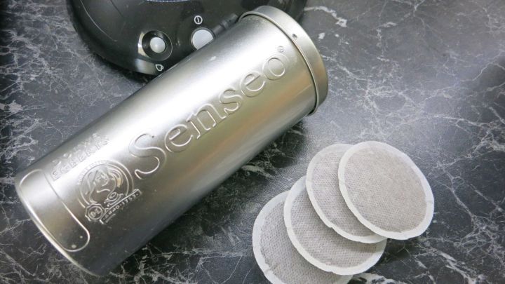 Ein silberner Beh&auml;lter f&uuml;r Senseo-Pads steht neben vier Pads auf einer schwarzen Arbeitsfl&auml;che, w&auml;hrend eine Kaffeemaschine im Hintergrund sichtbar ist.