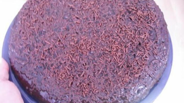 Ein runder Schokoladen-Brownie auf einem blauen Teller wird von einer Hand gehalten; er ist mit Schokostreuseln bedeckt.