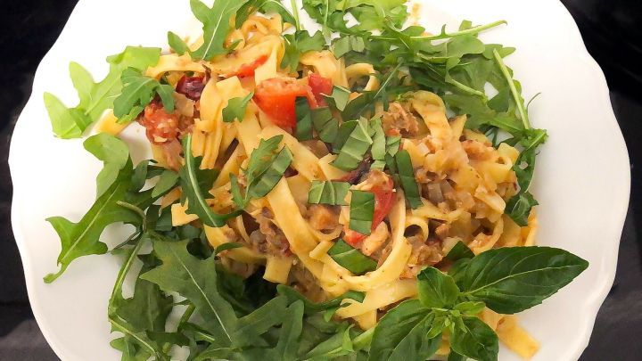 Nudelsalat liegt auf einem wei&szlig;en Teller, umgeben von frischem Rucola und Kr&auml;utern, mit Paprikast&uuml;cken und gehacktem Basilikum.