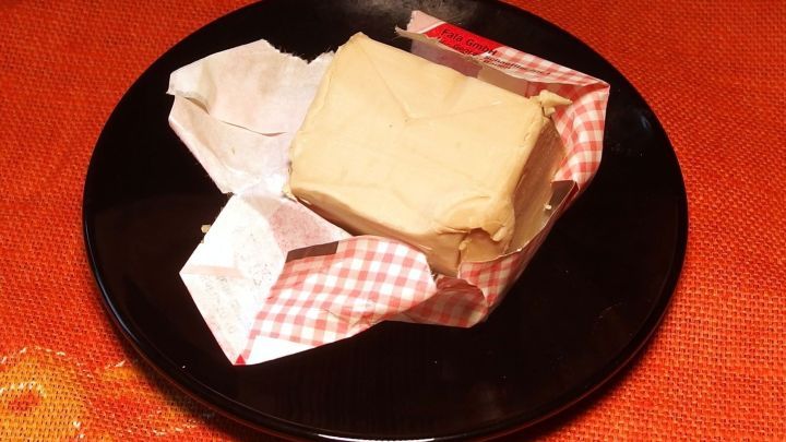 Ein St&uuml;ck Butter liegt teilweise verpackt auf einem schwarzen Teller, umgeben von einem orangen Hintergrund.