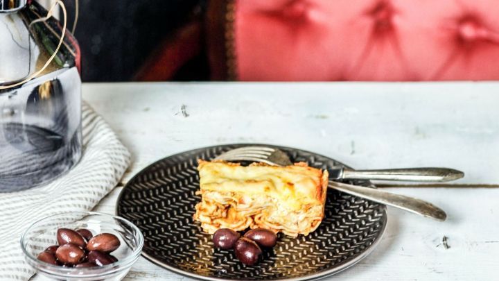 Eine appetitliche Lasagne auf einem kunstvollen Teller liegt auf einem Tisch, umgeben von einer Vase mit Trockenblumen und einem roten Stuhl.