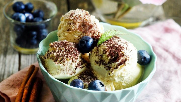 Eine Sch&uuml;ssel mit leckerem Dessert-Eis, dekoriert mit Blaubeeren, steht auf einem Holzuntergrund neben einer Vase mit Pfingstrosen.