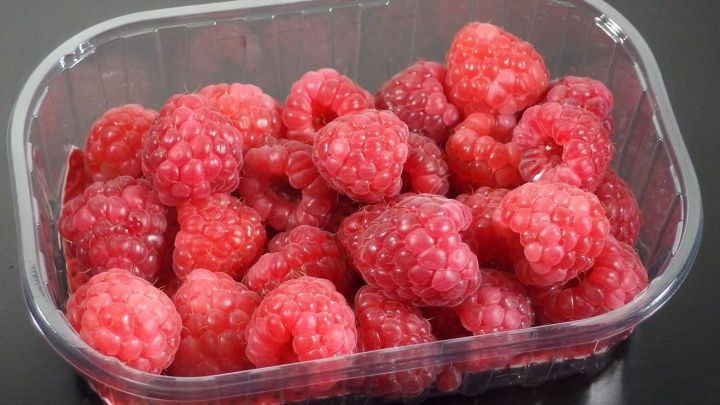 Frische Himbeeren liegen in einer transparenten Plastikschale auf einer dunklen Oberfl&auml;che, bereit zum Verzehr.