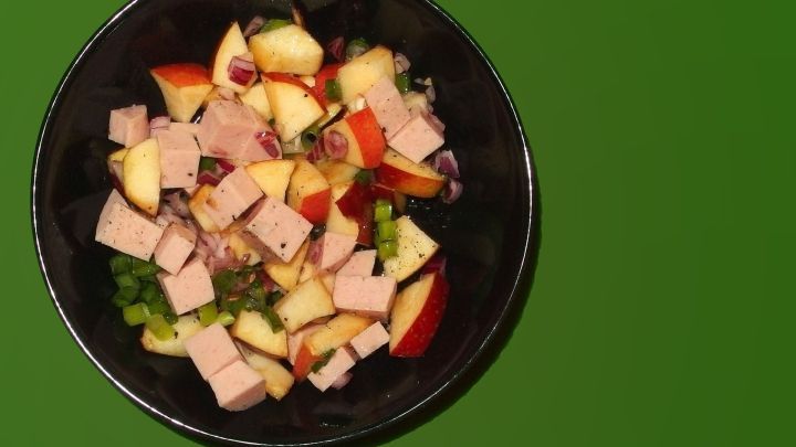 Wurstsalat mit Apfelst&uuml;cken und Zwiebeln in einer schwarzen Sch&uuml;ssel auf gr&uuml;nem Hintergrund.