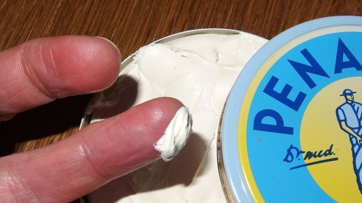 Eine Hand nimmt Penatencreme aus einer Dose, um trockene Lippen zu behandeln, auf einem holzfarbenen Tisch.