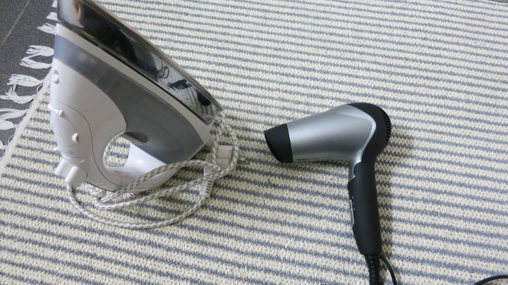 Ein B&uuml;geleisen liegt auf einem Fu&szlig;bodenbelag, w&auml;hrend daneben ein F&ouml;n steht. Beide Ger&auml;te sind bereit zum Einsatz.