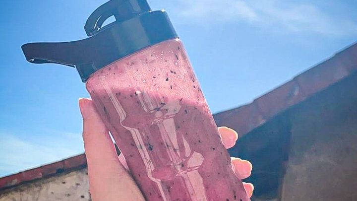 Ein Smoothie wird in der Hand gehalten, w&auml;hrend die Sonne am blauen Himmel scheint, umgeben von einem Geb&auml;udetr&auml;ger.