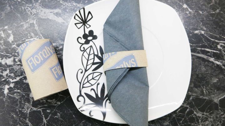 Ein eleganter Serviettenring aus Karton h&auml;lt eine gefaltete, graue Serviette auf einem stilvollen, dekorierten Teller, platziert auf einer dunklen Oberfl&auml;che.