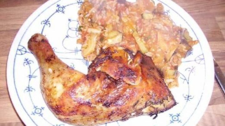 Ein gebratenes H&auml;hnchenbein liegt auf einem Teller neben Ratatouille mit Reis, serviert auf einem Holztisch.