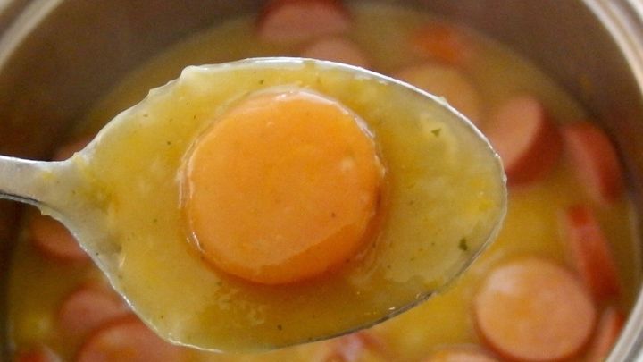 Ein L&ouml;ffel mit einem ganzen Eigelb hebt sich &uuml;ber einen k&ouml;chelnden M&ouml;hreneintopf, der mit W&uuml;rstchen garniert ist.