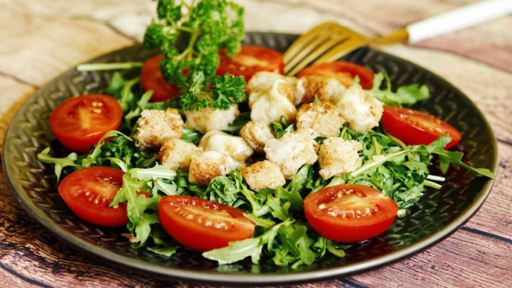 Ein frischer Salat mit knusprigen K&auml;sew&uuml;rfeln und roten Tomatenscheiben auf einem Teller, umgeben von gr&uuml;nen Bl&auml;ttern.