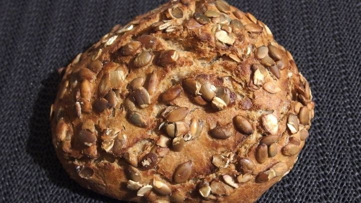 Ein frisch gebackenes Brot mit einer knusprigen Oberfl&auml;che liegt auf einem dunklen Stoff. Es hat eine nussige Farbe und Textur.