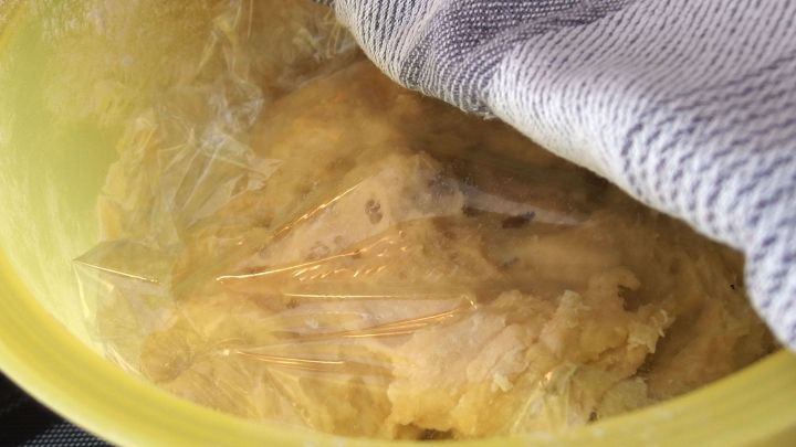 Ein gelber Beh&auml;lter enth&auml;lt Teig, der mit Folie abgedeckt und mit einem Tuch gesch&uuml;tzt ist, um ein Austrocknen zu verhindern.