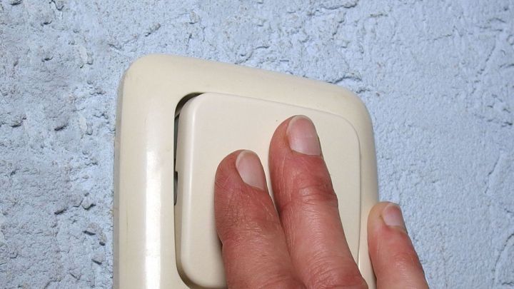 Eine Hand dr&uuml;ckt einen Lichtschalter an einer hellblauen Wand, um das Licht auszuschalten.