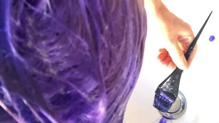 Ein Kopf mit lila gef&auml;rbtem Haar wird mit einem Pinsel gef&auml;rbt, w&auml;hrend Farbreste im Waschbecken sichtbar sind.