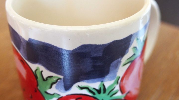 Ein bunter Porzellanbecher steht auf einem Tisch, seine bemalte Oberfl&auml;che zeigt rote Tomaten und gr&uuml;ne Bl&auml;tter.