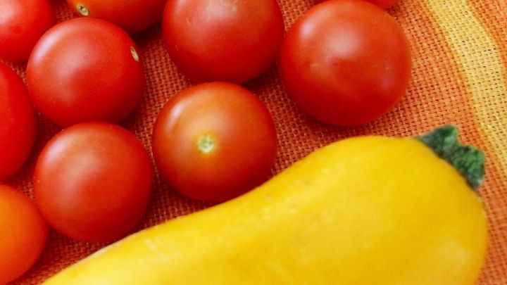 Eine gelbe Zucchini liegt neben einer Ansammlung von roten Tomaten auf einem orangefarbenen Tuch.