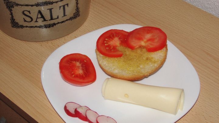 Ein Br&ouml;tchen mit Oliven&ouml;l, belegt mit Tomatenscheiben und K&auml;se, liegt auf einem Teller neben Radieschen.