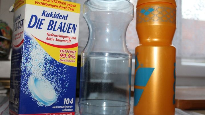 Eine Wasserflasche steht neben einem orangefarbenen Beh&auml;lter und einer Reinigungsmittelpackung auf einer Tischfl&auml;che.