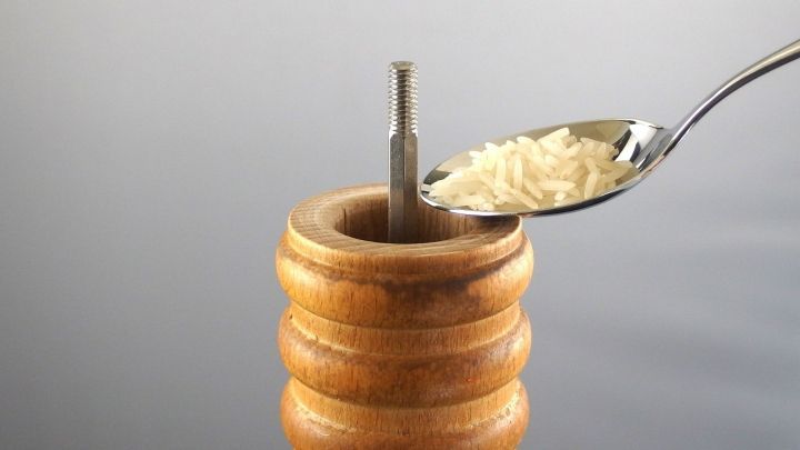Eine Holz-Pfefferm&uuml;hle wird mit einer L&ouml;ffelportion Reis gef&uuml;llt, vor einem neutralen Hintergrund sichtbar.