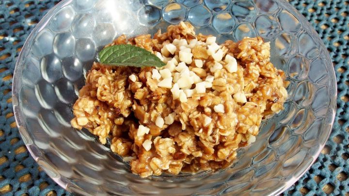 Gebratene Haferflocken liegen in einer glasigen Schale, garniert mit N&uuml;ssen und einem Minzblatt, vor einem strukturierten Hintergrund.