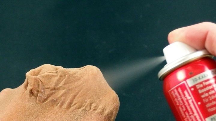 Eine Hand spr&uuml;ht Haarspray auf eine Laufmasche in einer Strumpfhose, um sie zu reparieren. Der Hintergrund ist schwarz.