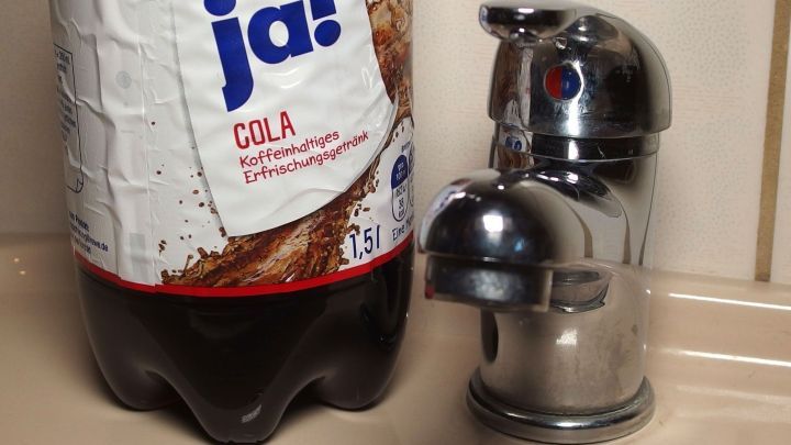 Ein gl&auml;nzender Wasserhahn steht neben einer Cola-Flasche, w&auml;hrend Cola in das Sp&uuml;lbecken flie&szlig;t.