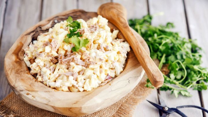 Eine Schale mit cremigem Thunfischsalat steht auf einem Holztisch, umgeben von frischen Kr&auml;utern und einem Holzl&ouml;ffel.
