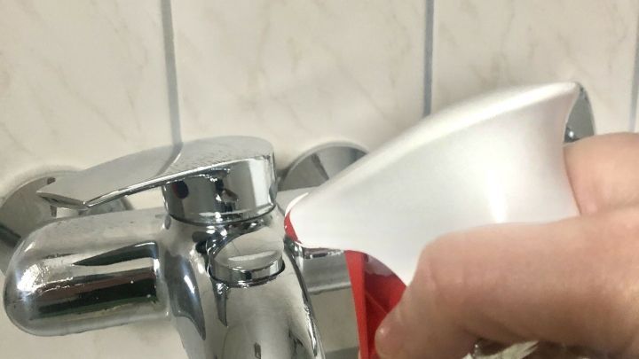 Ein Handgriff spr&uuml;ht Glasreiniger auf einen Wasserhahn in einem Badezimmer mit wei&szlig;en Fliesen.