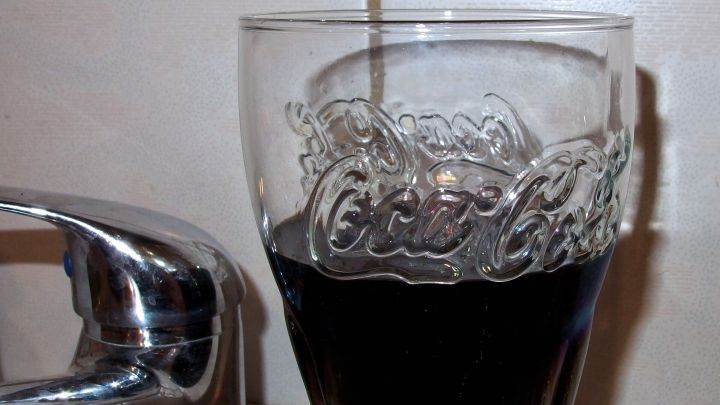 Ein Glas Cola steht neben einem Wasserhahn, w&auml;hrend Cola langsam hineingegossen wird, um den Abfluss zu reinigen.