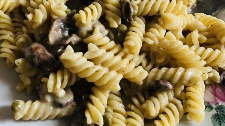 Ein Nudelsalat mit spiraligen Nudeln, Pilzen und einer cremigen Sauce, serviert auf einem wei&szlig;en Teller vor einem Holzuntergrund.