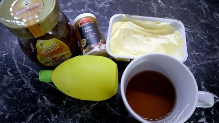 Ein Becher Tee steht auf einer Marmoroberfl&auml;che, umgeben von Honig, Butter, einer Zitrone und Gew&uuml;rzen, um Halsschmerzen zu lindern.