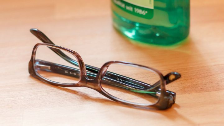 Eine Brille liegt auf einem Holzuntergrund, w&auml;hrend ein Reinigungsmittel daneben steht &ndash; ideal f&uuml;r die Pflege unterwegs.