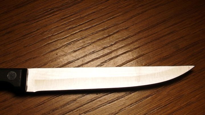 Ein scharfes Messer liegt still auf einer h&ouml;lzernen, leicht strukturierten Tischoberfl&auml;che, kontrastreich beleuchtet.