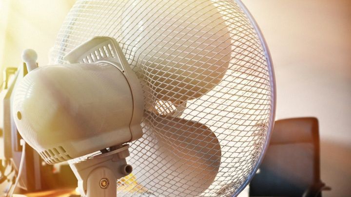 Ventilator dreht sich in einem B&uuml;roraum und sorgt f&uuml;r Luftzirkulation. Sunschein beleuchtet den Raum hinter dem Ger&auml;t.